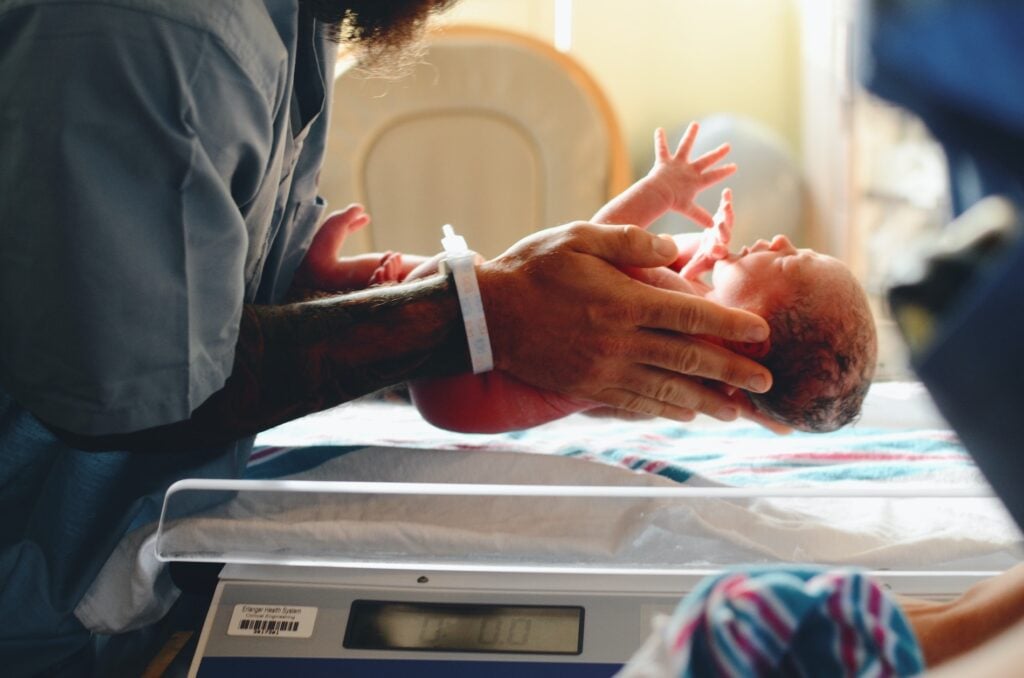 Nouveau-né posé sur une balance médicale, soutenu par un professionnel illustrant le rôle sage-femme dans l’accompagnement des premières heures de vie.