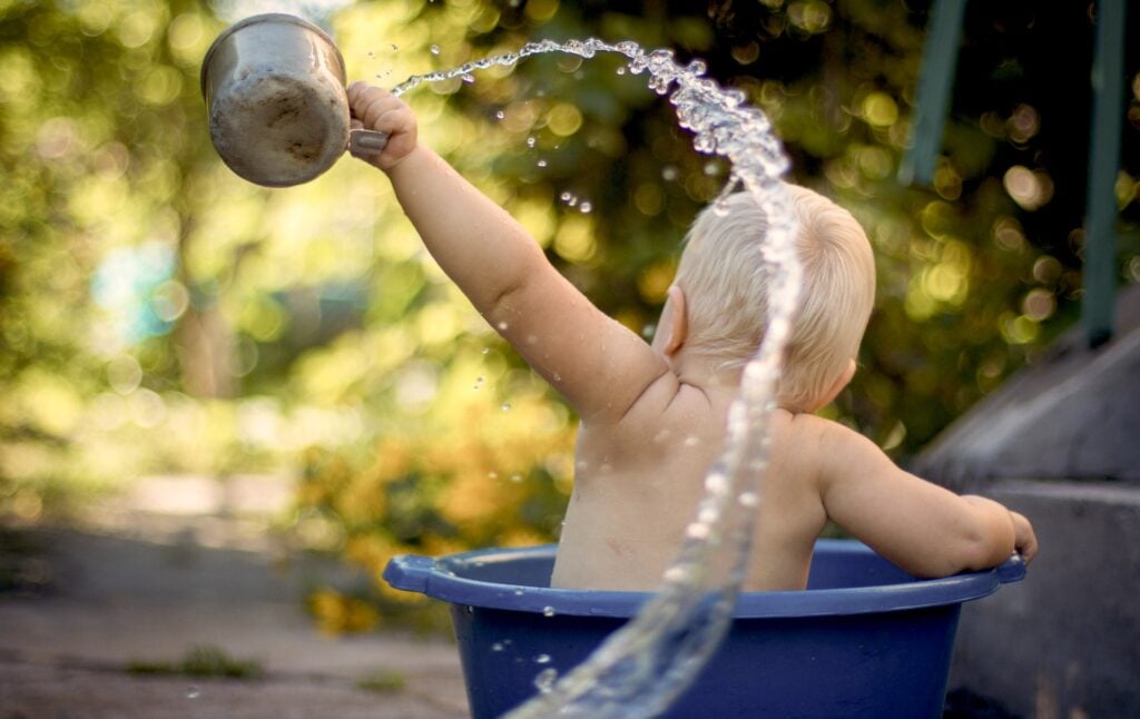 Bébé blond assis dans une bassine en extérieur et versant de l’eau avec une casserole, jeu d’eau ludique en été