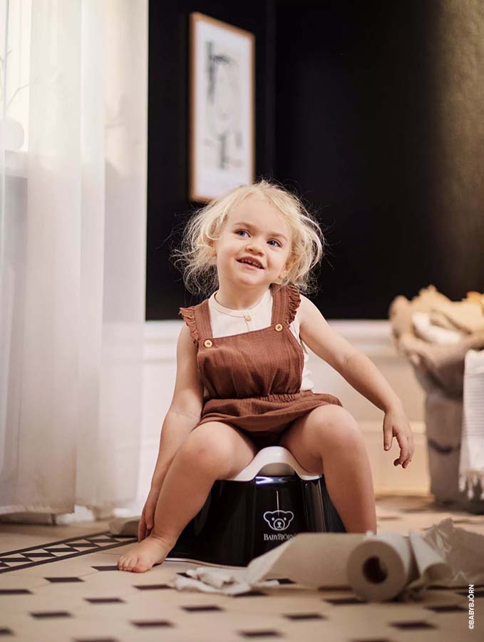 Petite fille assise sur un pot BabyBjörn noir dans une salle de bain élégante, avec du papier toilette sur le sol. Signes que bébé est prêt pour le pot
