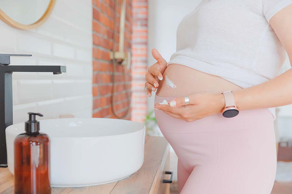 Photo d'une femme enceinte dans une salle de bain lumineuse, appliquant délicatement une huile naturelle sur son ventre arrondi pour prévenir les vergetures. À côté d'elle, sur le plan de travail, se trouve un flacon-pompe en verre ambré contenant l'huile. L'image reflète l'importance des soins de la peau naturels et efficaces pour les futures mamans.