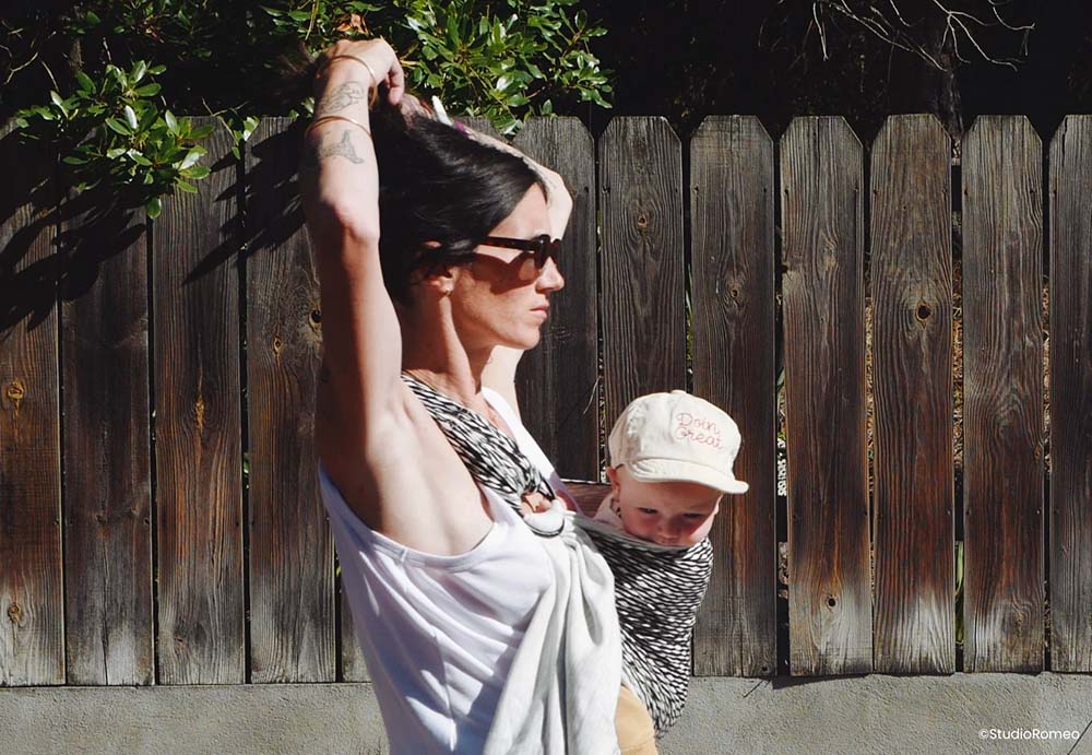Une mère au look moderne, avec des tatouages visibles sur le bras et portant des lunettes de soleil, marche au soleil près d'une clôture en bois. Elle porte fièrement son bébé, vêtu d'un chapeau inscrit "Born to be Great", dans un porte-bébé rayé contre sa poitrine. Leur complicité et leur proximité transparaissent, malgré l'absence de famille à proximité. L'image capture la force et la résilience de la maternité en solo, montrant comment l'amour maternel peut transcender les distances et les défis.