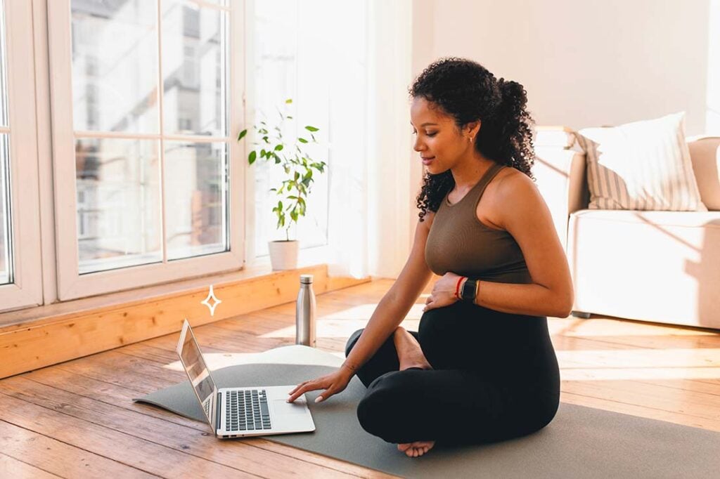 Femme enceinte assise en tailleur devant un ordinateur sur un tapis de sport, séance de préparation prénatale en ligne