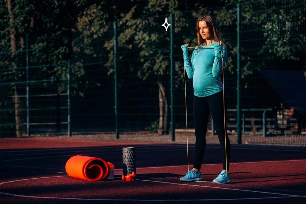 Femme enceinte s’entraînant avec élastiques sur terrain de sport extérieur, activité physique adaptée à la grossesse