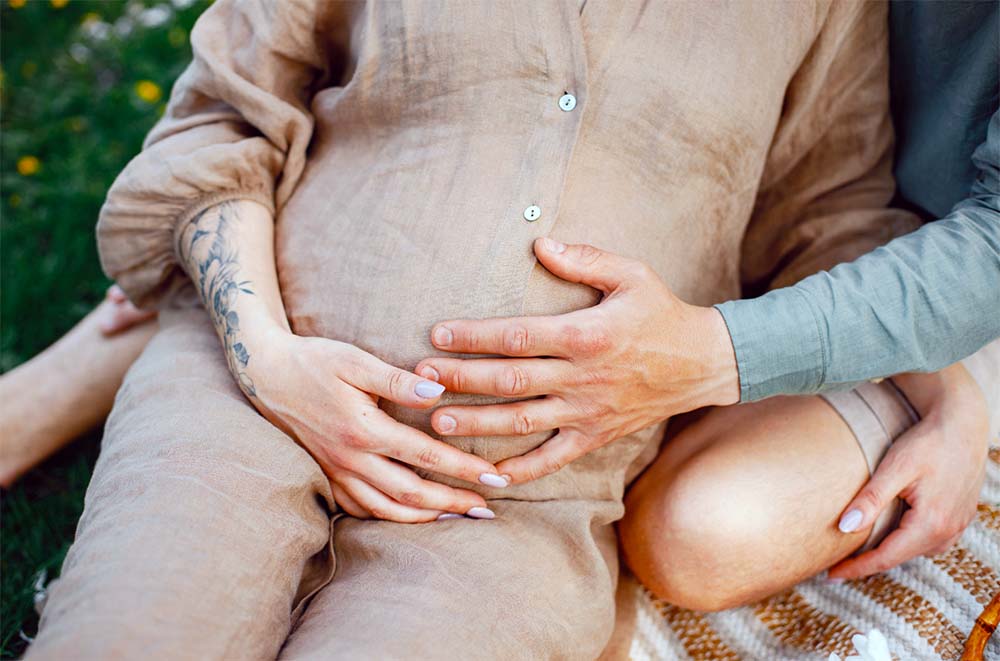 Partage de moments de complicité en couple, mains entrelacées sur un ventre de femme enceinte, symbolisant l'union et le soutien dans l'attente de l'accouchement naturel.