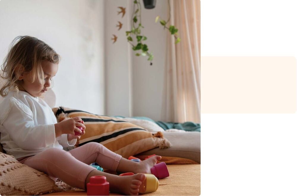 Période d’adaptation crèche bébé, fillette assise sur un lit jouant calmement avec des blocs colorés dans une ambiance douce et rassurante