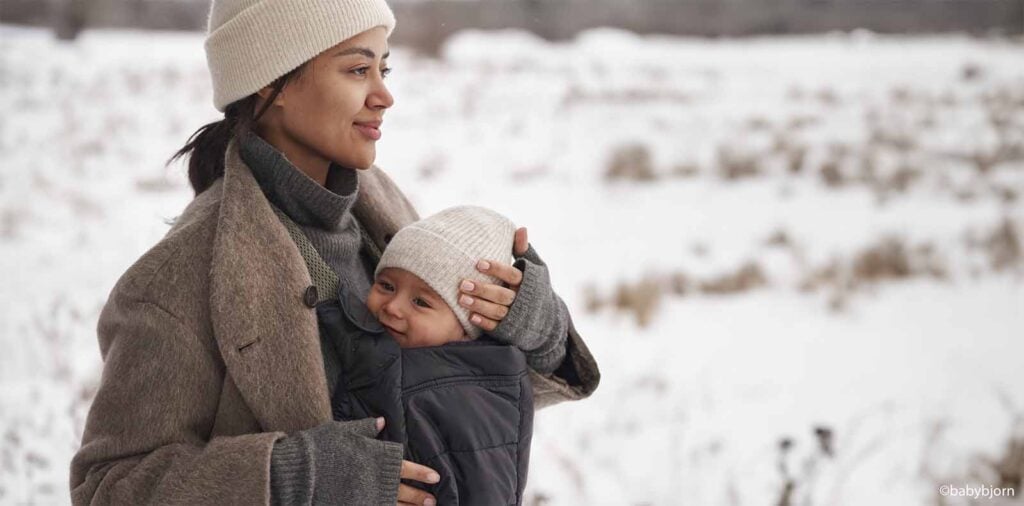 Maman portant son bébé en porte-bébé dans la neige avec bonnets et vêtements chauds d'hiver