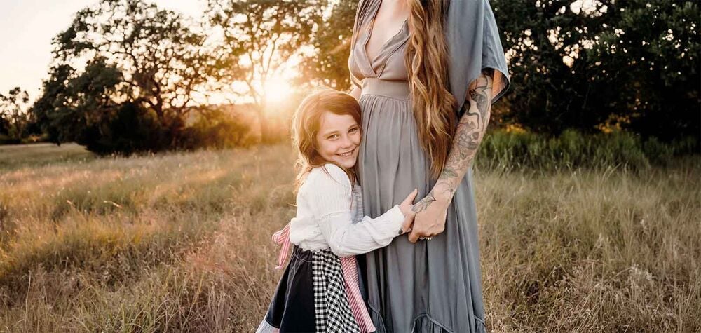 Maman enceinte avec fillette souriante dans un champ au coucher du soleil, couple en famille recomposée attendant un bébé, grossesse et fratrie