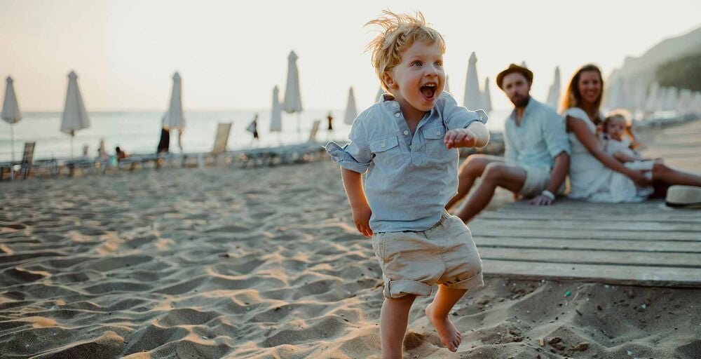 Petit garçon courant sur le sable, parents assis avec un bébé en arrière-plan, préparer l'arrivée d'un bébé en famille recomposée, rassurer les enfants