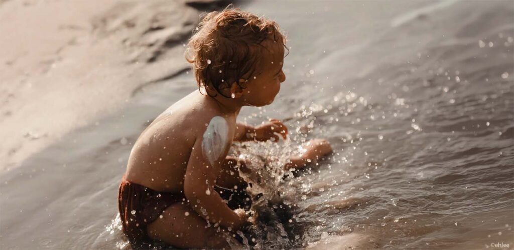 Bébé jouant dans l'eau avec de la crème solaire visible sur le dos, protection bébé soleil à la plage, bien appliquer l'écran solaire