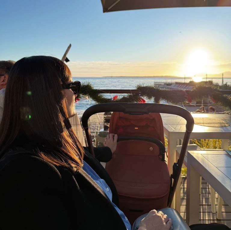 Maman avec poussette Maxi-Cosi face au coucher de soleil en bord de mer, quelle poussette voyage choisir pour les vacances avec bébé